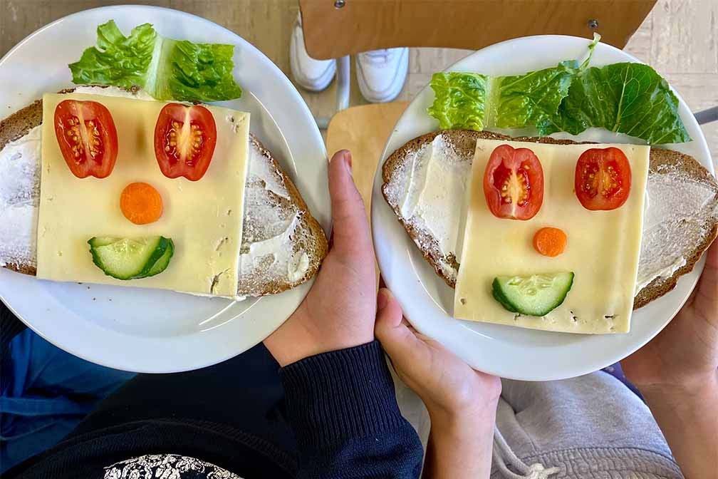 Zwei Kinder halten ihren Teller mit belegten Broten. Die Brote sind so mit Gemüse und Käse belegt das sie aussehen wie Gesichter