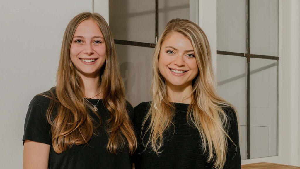 Zwei blonde Frauen die frontal zur Kamera stehen, lächeln und sich seitlich im Arm halten