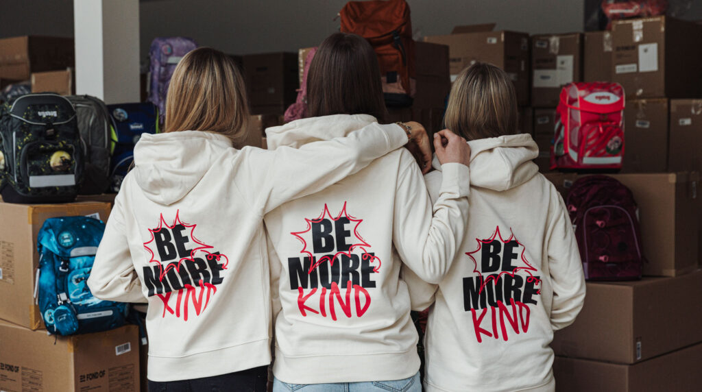 3 Frauen die mit dem Rücken zur Fotografin stehen. Auf den Pullis die sie anhaben steht Be more Kind